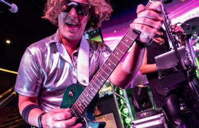 Man with curly hair and sunglasses playing electric guitar on stage under colorful lights.
