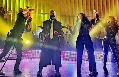 Four singers performing energetically on stage with colorful spotlights behind them.
