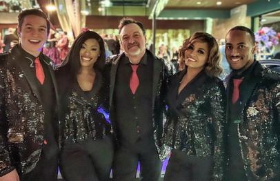 Five people in matching black sequin suits with red ties smiling at an event.