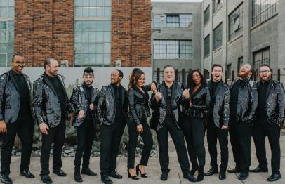 Nine people in black jackets smiling and posing together outdoors in an urban setting.