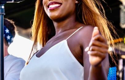Smiling woman singing on stage in bright sunlight, wearing a white top.
