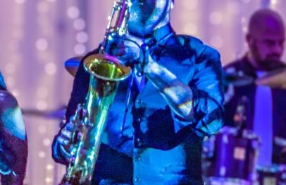 Musician playing saxophone on stage with colorful lights and a drummer in the background.