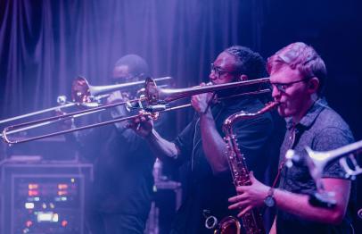 Three musicians play trombone and saxophone on stage under purple lighting.