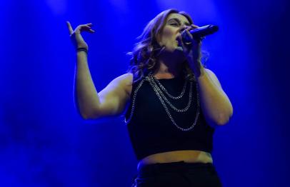 Woman singing on stage under blue lights, wearing a black top and silver chains.
