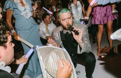 Man with green hair singing into a microphone at a lively party with people around him.