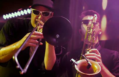 Two musicians wearing sunglasses play trumpet and saxophone under purple stage lights.