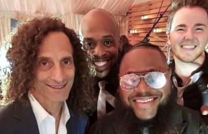 Four smiling men in suits posing for a selfie at an indoor event.