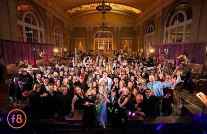 Large group of people in formal attire posing in a grand ballroom with arched windows.