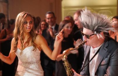 Bride dancing beside a man with spiky gray hair playing saxophone at a lively party.