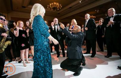 Man proposing on one knee to a woman in a blue dress at a formal event, surrounded by people.