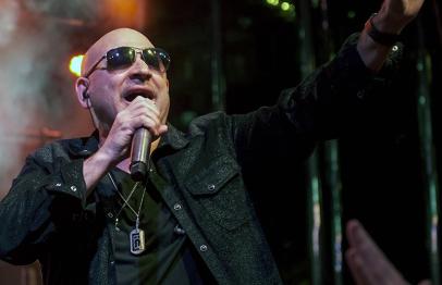 Singer in sunglasses and black jacket performing on stage, hand raised, colorful lights behind.