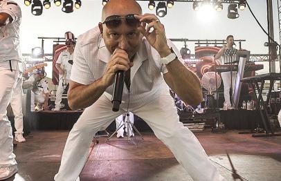 Man in white performing energetically on stage with band members in the background.