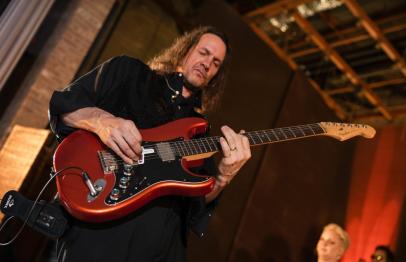Man playing a red electric guitar on stage under warm lighting.