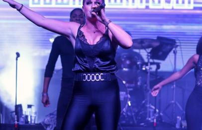Female singer performing energetically on stage with purple lighting and background display.