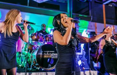 Three female vocalists and a live band performing on a colorful stage.