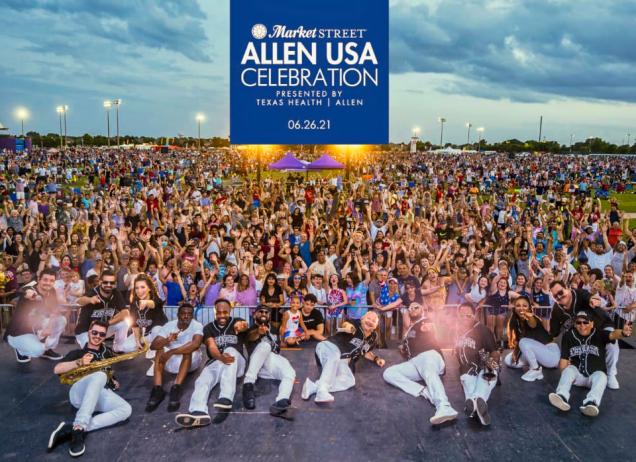 Large crowd at an outdoor festival with musicians posing on stage at dusk.