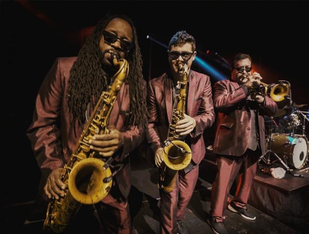 Three musicians in matching suits play saxophones and trumpet on stage.