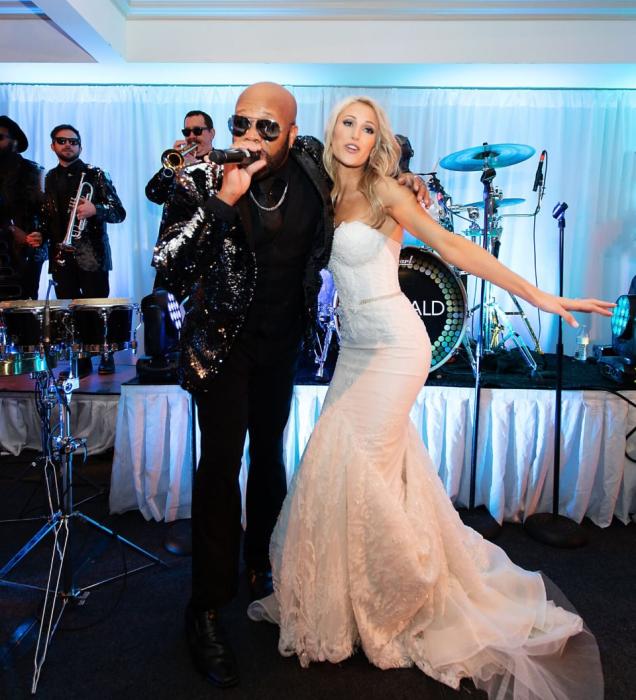 Bride in a white gown poses with a singing performer on stage with a live band.
