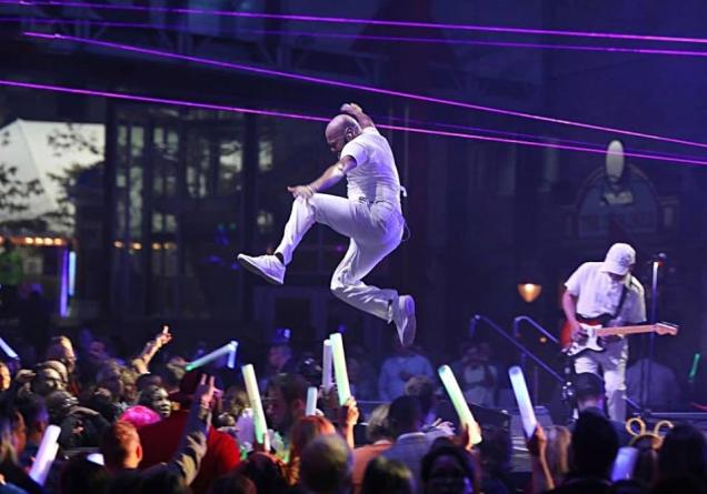 Performer in mid-air jump on stage with guitarist, crowd holding glow sticks.