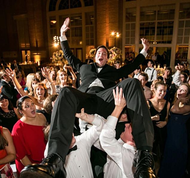 Man in a tuxedo lifted and cheered by a lively crowd at a formal event.