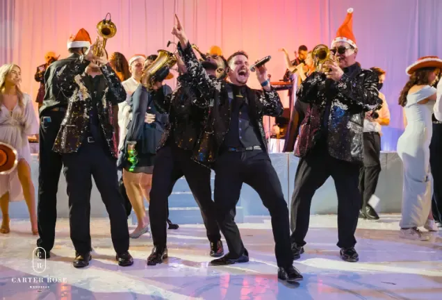 Band in festive attire performing energetically on stage, some wearing Santa hats.