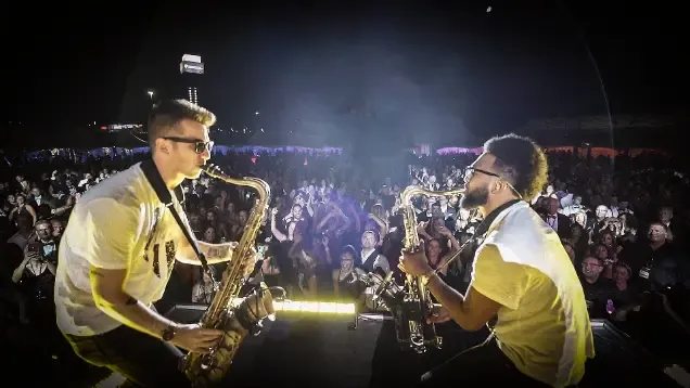 Two musicians playing saxophones on stage before a cheering crowd at night.