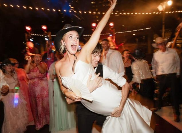 Bride in a cowboy hat being lifted on a lively dance floor at a wedding reception.