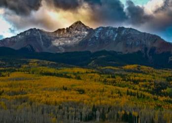 Snow-capped mountain at sunset above a forest with vibrant yellow autumn trees.
