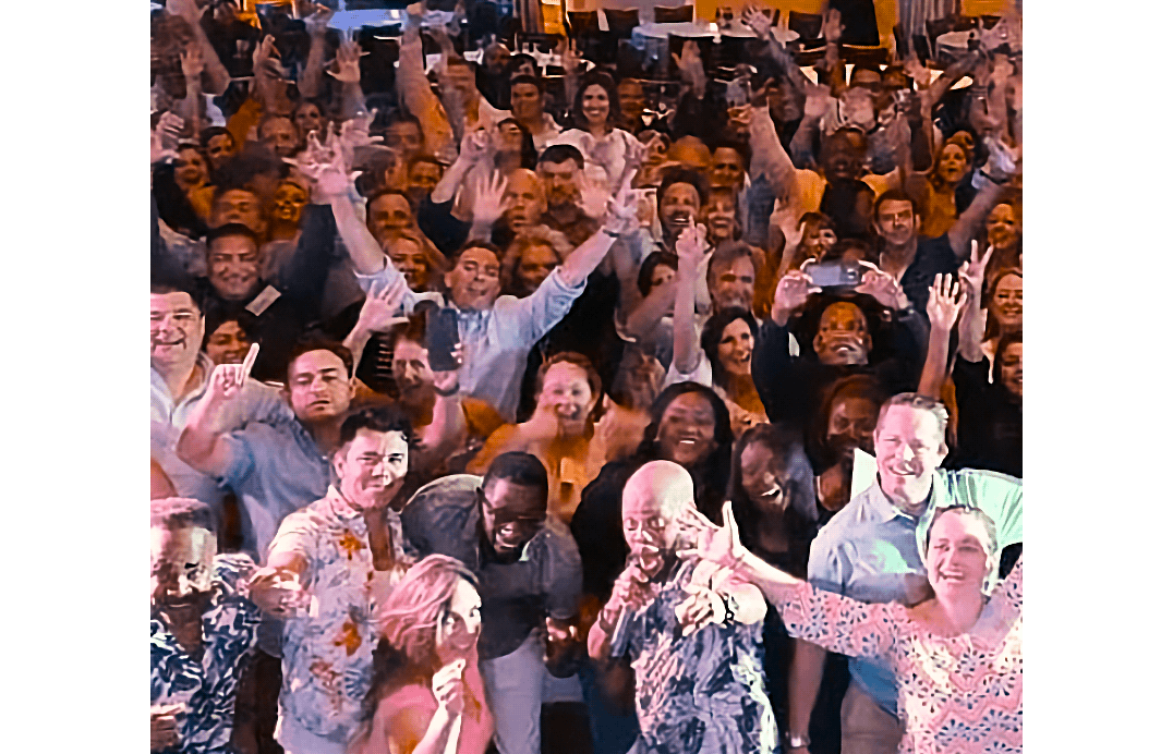 Large group of people smiling and waving at a lively indoor event.