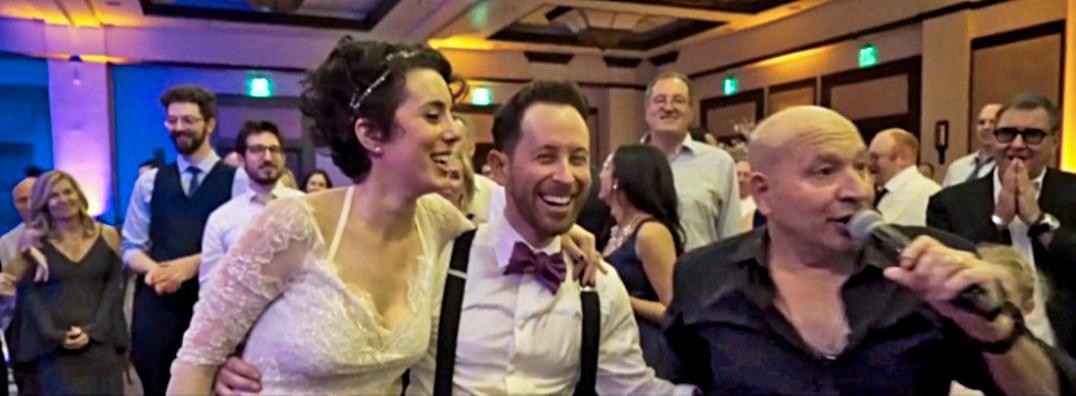 Bride and groom laughing with a singer at a lively wedding reception.