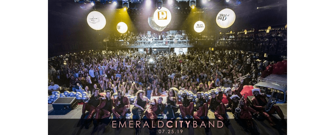 Large crowd with a band posing on stage under bright concert lights.