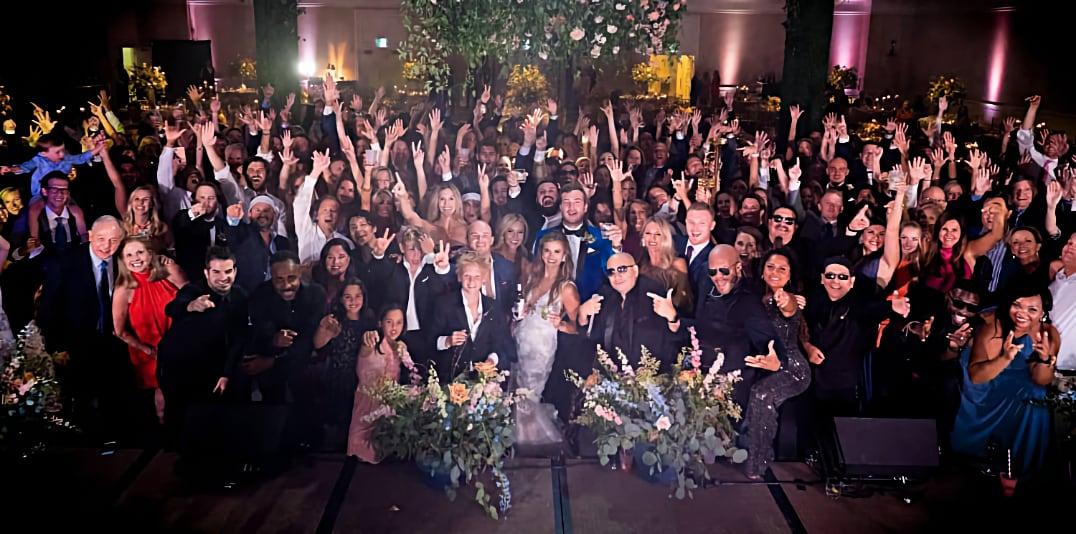 Large group of people celebrating at a wedding reception, raising hands and smiling.