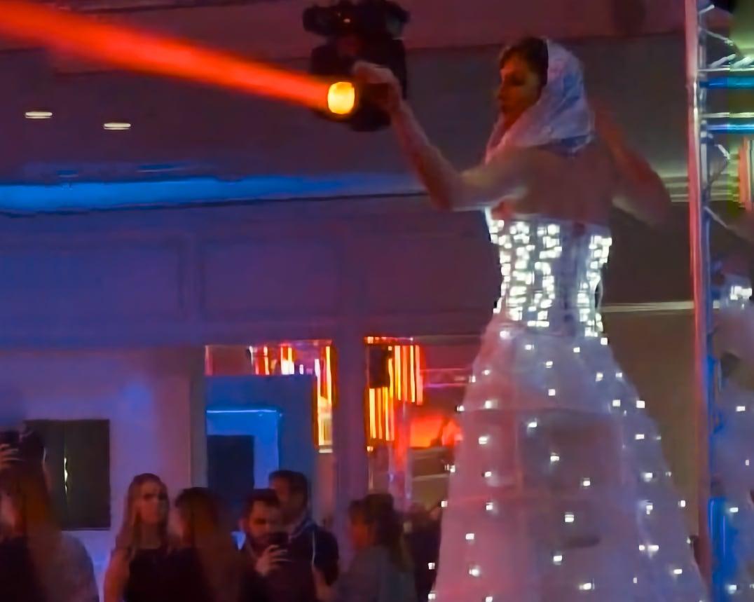 Performer in a glowing LED dress shines a spotlight over a crowd at an indoor event.