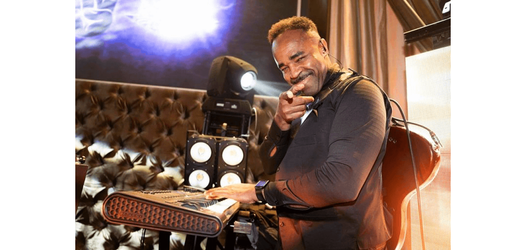 DJ smiling and pointing while playing music at a club with stage lights behind him.