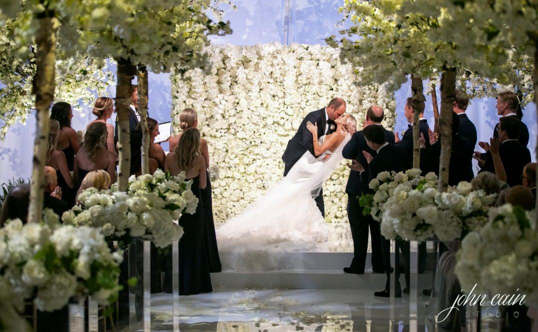 Bride and groom kiss at flower-filled wedding ceremony, surrounded by applauding guests.