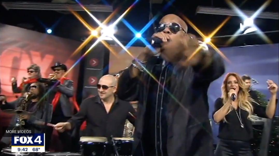 Band performing live on TV set, lead singer reaching forward, bright stage lights shine.