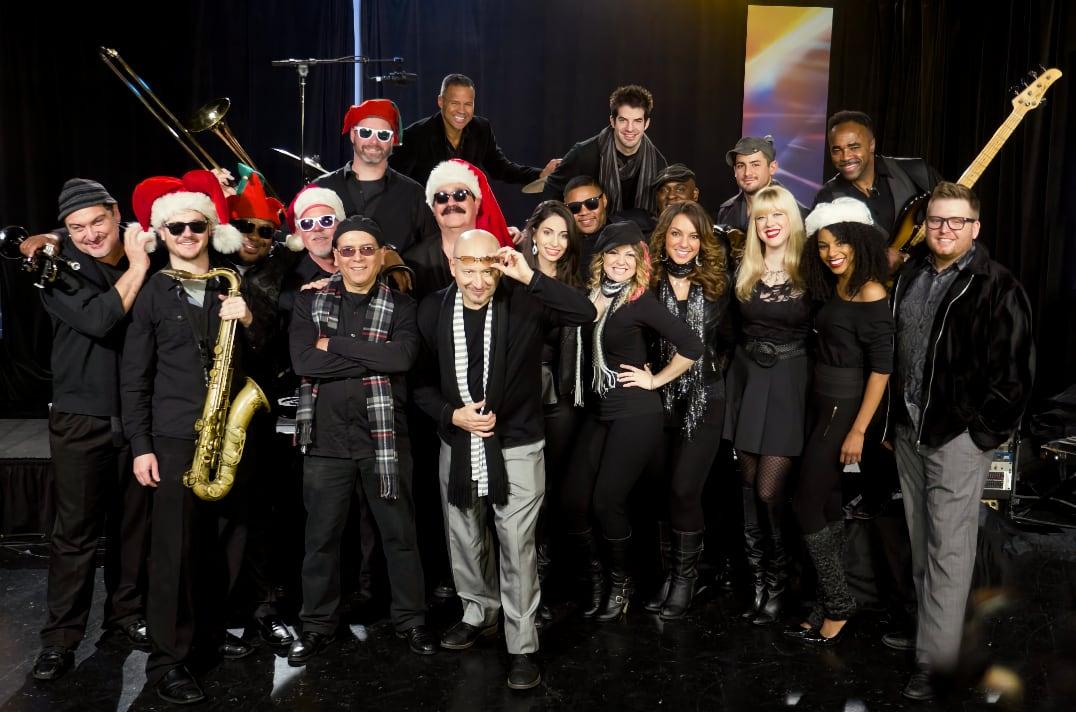 Band members in black outfits and Santa hats pose together on stage.