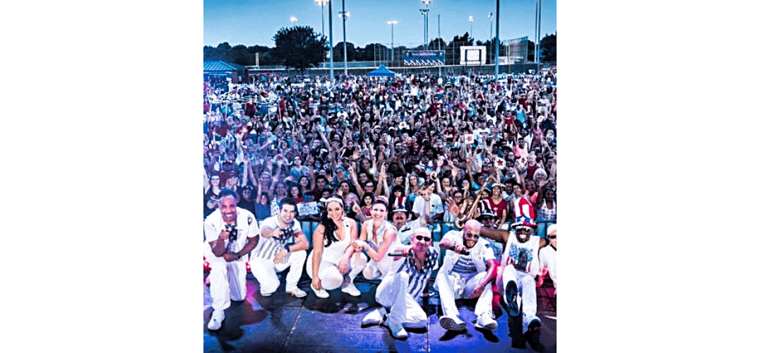 Performers in white outfits pose on stage with a large, cheering crowd behind them outdoors.