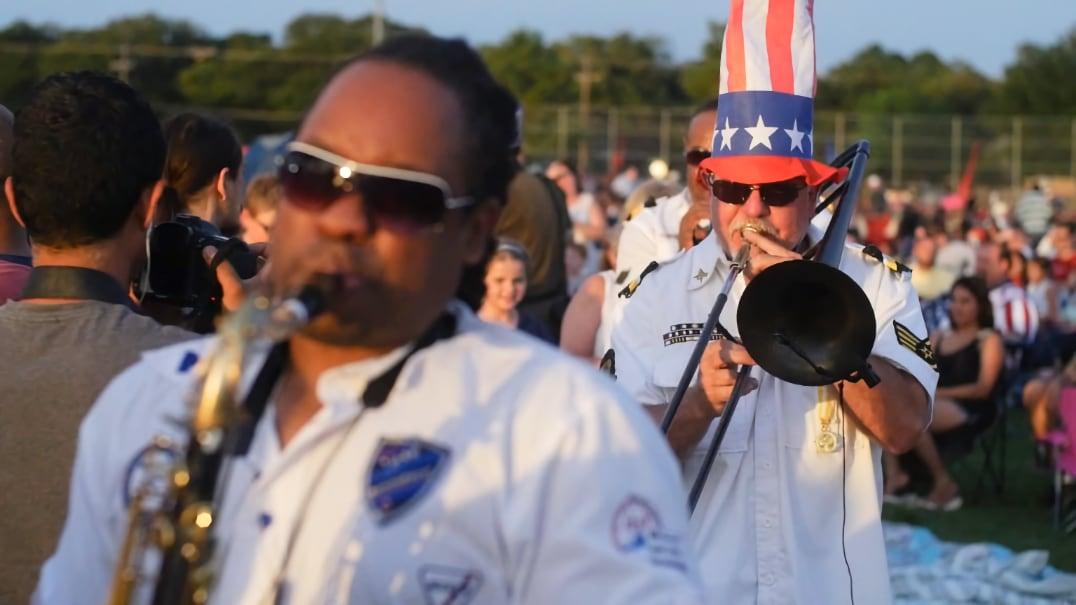 Musicians play brass instruments outdoors, one wearing a tall American flag hat.