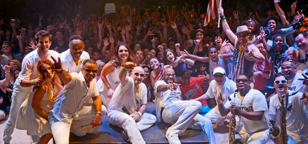 Musicians in white posing on stage with an enthusiastic crowd in the background.