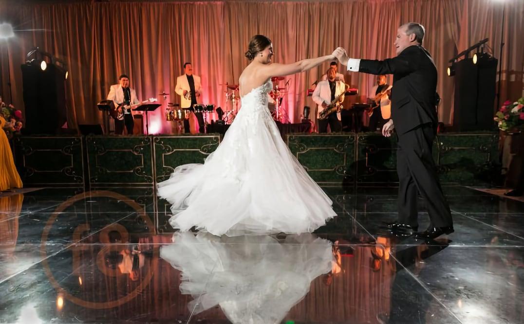 Bride and father dancing on a reflective floor with a live band in the background.