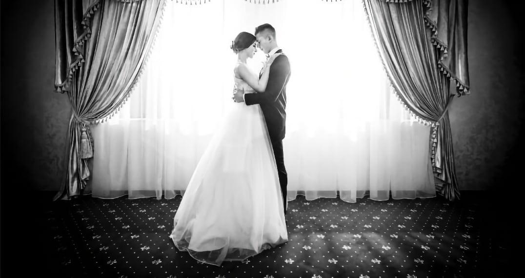Bride and groom embrace in front of a bright window with elegant curtains.