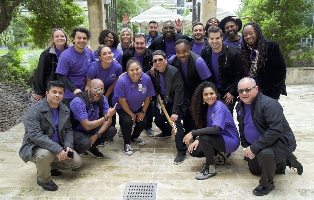 Group of people smiling outdoors, most wearing purple shirts, some in black jackets.