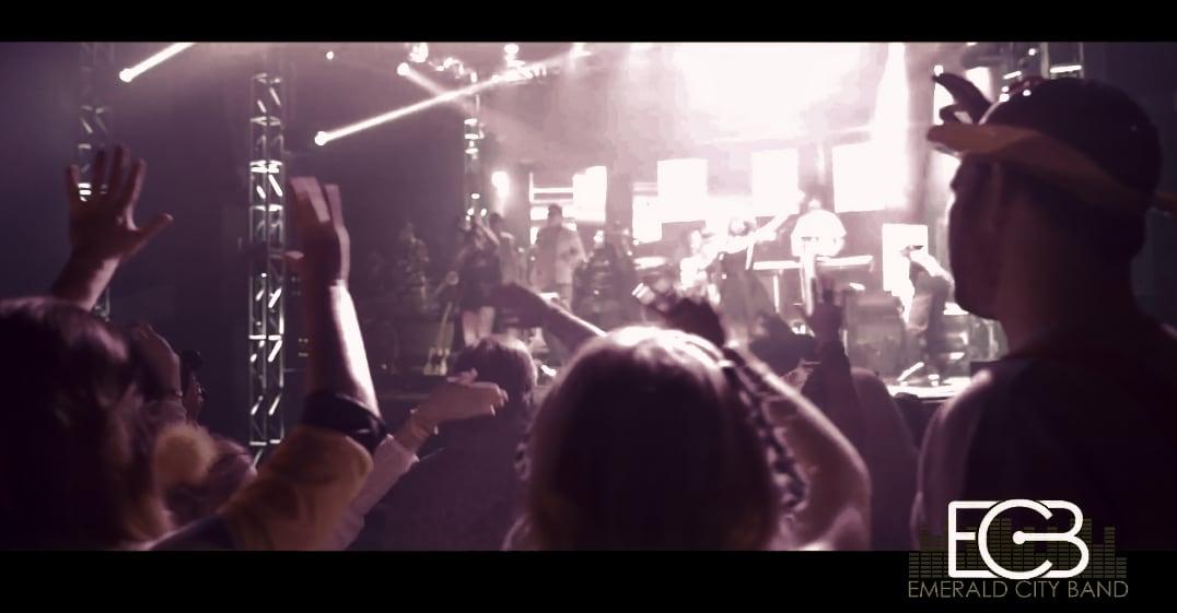 Concert crowd raising hands under bright stage lights, people silhouetted in the foreground.