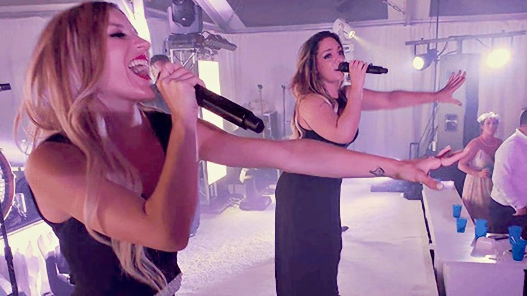 Two women in black dresses singing passionately on stage with microphones.
