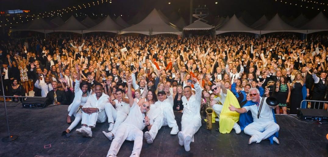 Performers in white pose on stage before a cheering crowd at a nighttime outdoor event.