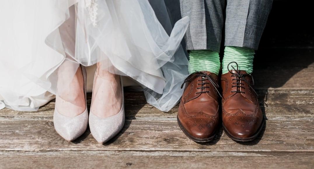 Bride’s white shoes beside groom’s brown shoes and green socks on wooden floor.
